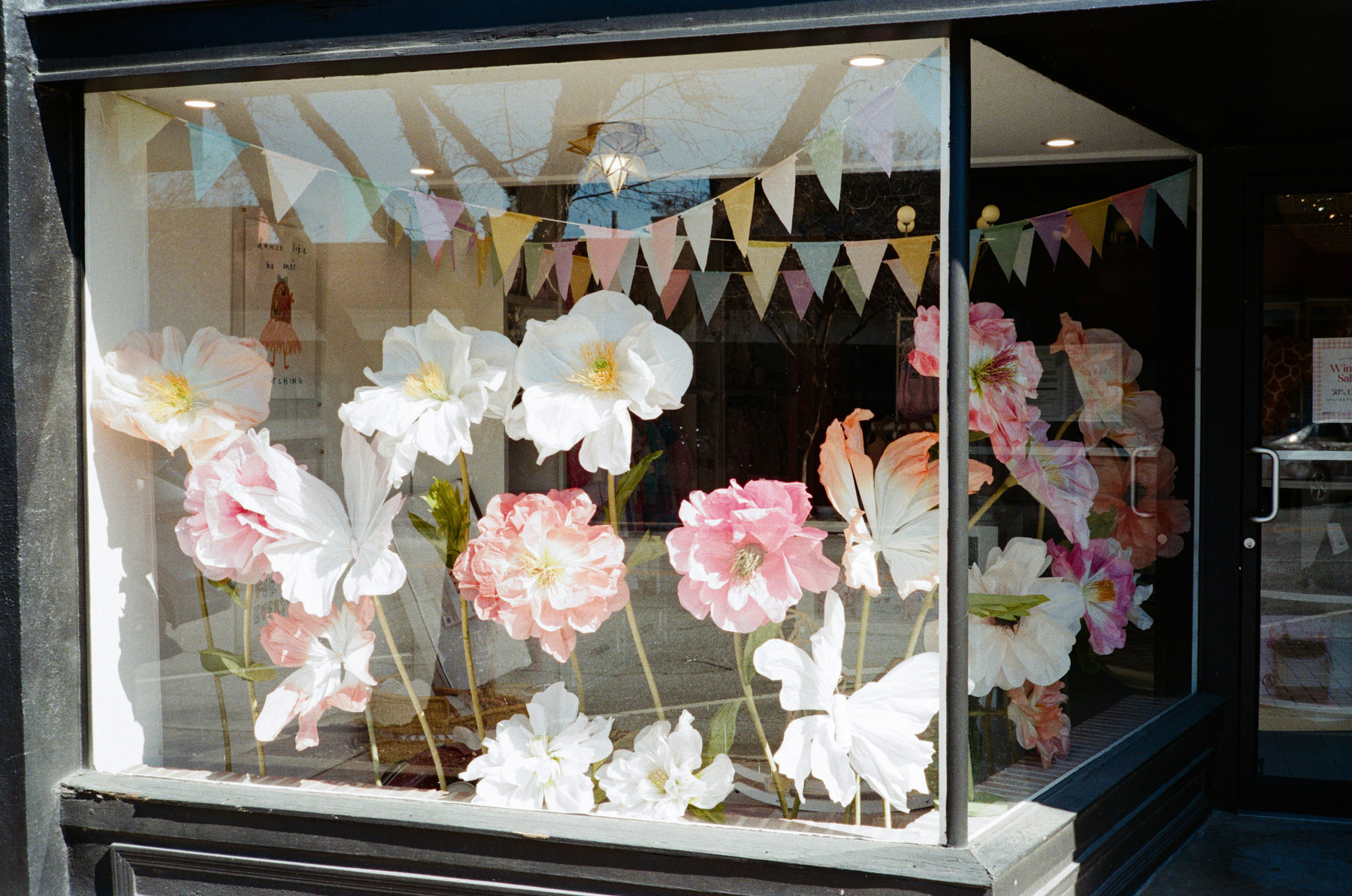 Auto-generated description: A storefront display features large paper flowers in pastel colors with a triangular flag banner above.