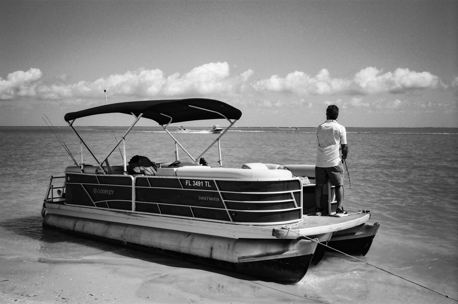 Auto-generated description: A man stands on a pontoon boat at the water&rsquo;s edge, looking out over a calm sea under a sky with scattered clouds.
