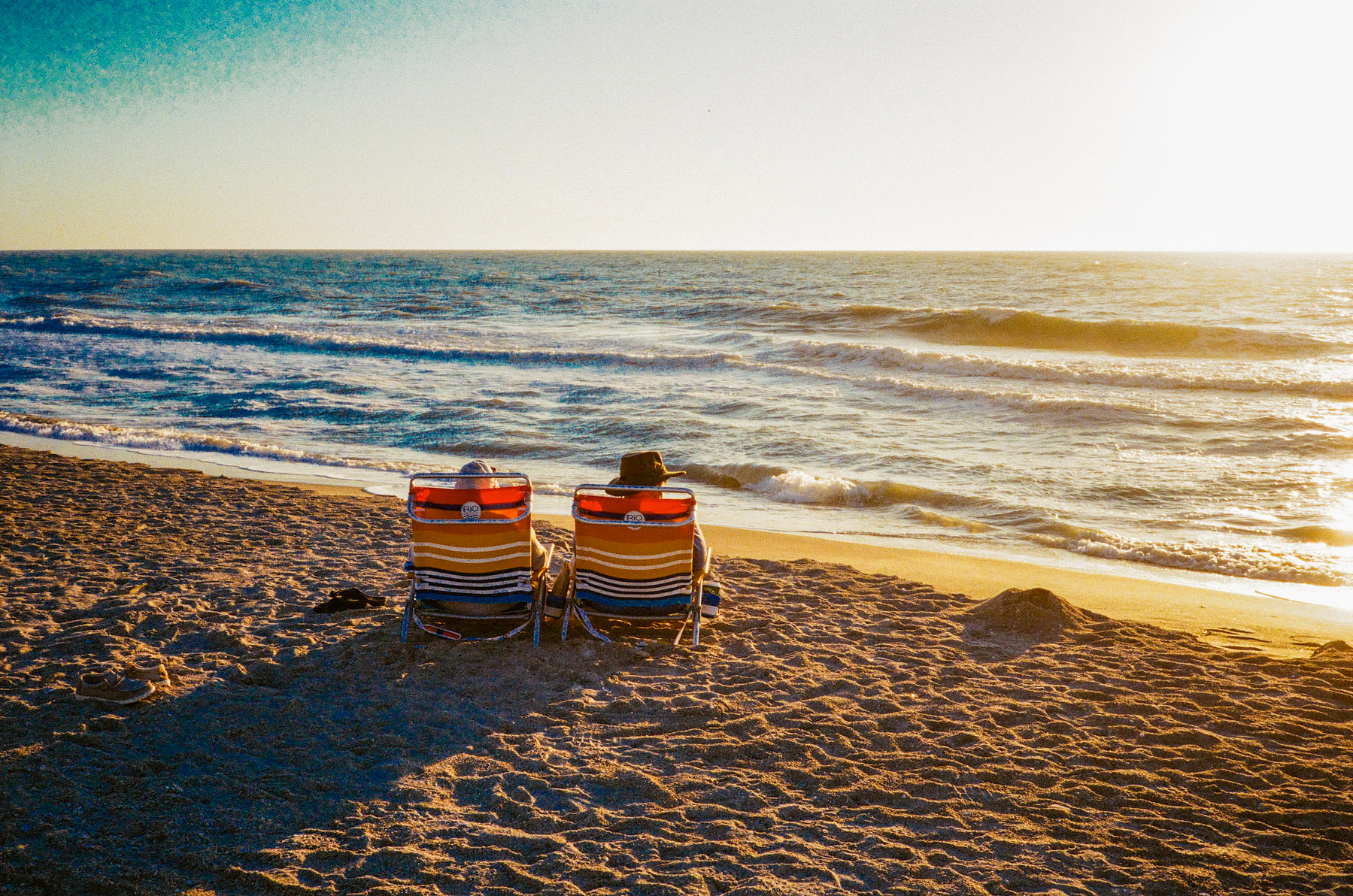 Auto-generated description: Two people sit in colorful striped beach chairs on the sand, facing the ocean at sunset.