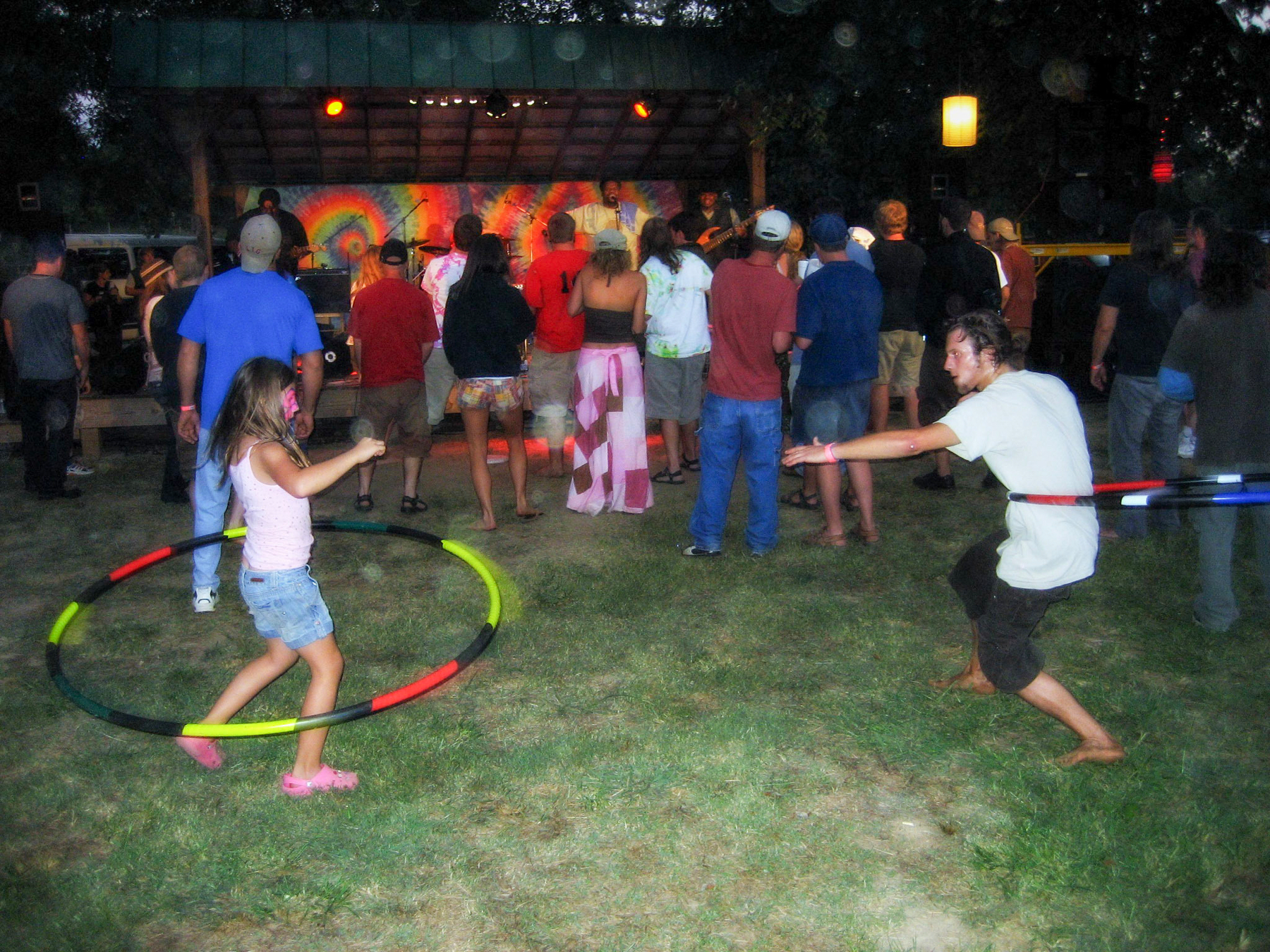 Auto-generated description: A girl and a man are hula-hooping on a grassy area in front of a crowd watching a band perform on stage.