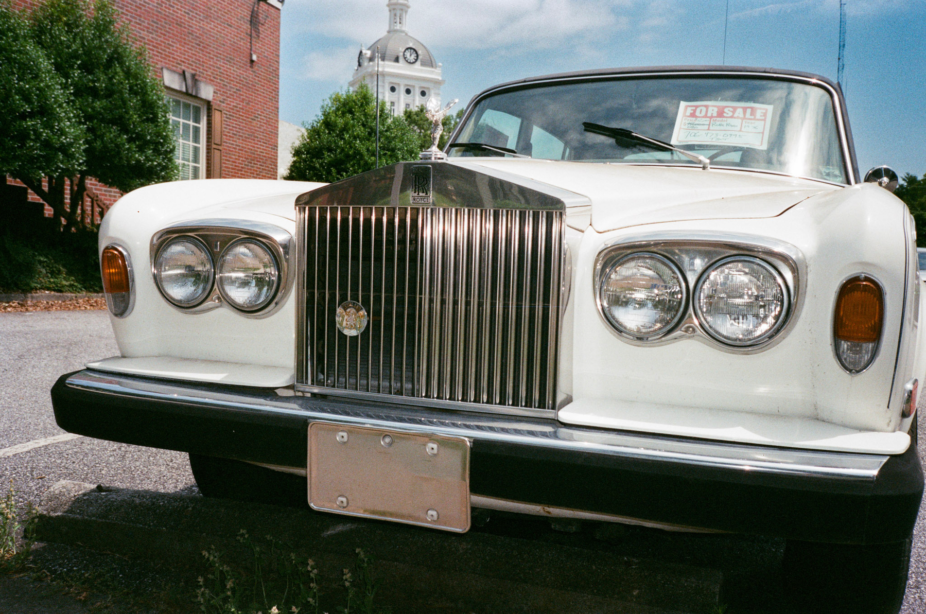 Auto-generated description: A vintage white Rolls-Royce is parked on a street with a For Sale sign in its window, framed by trees and a classic building in the background.