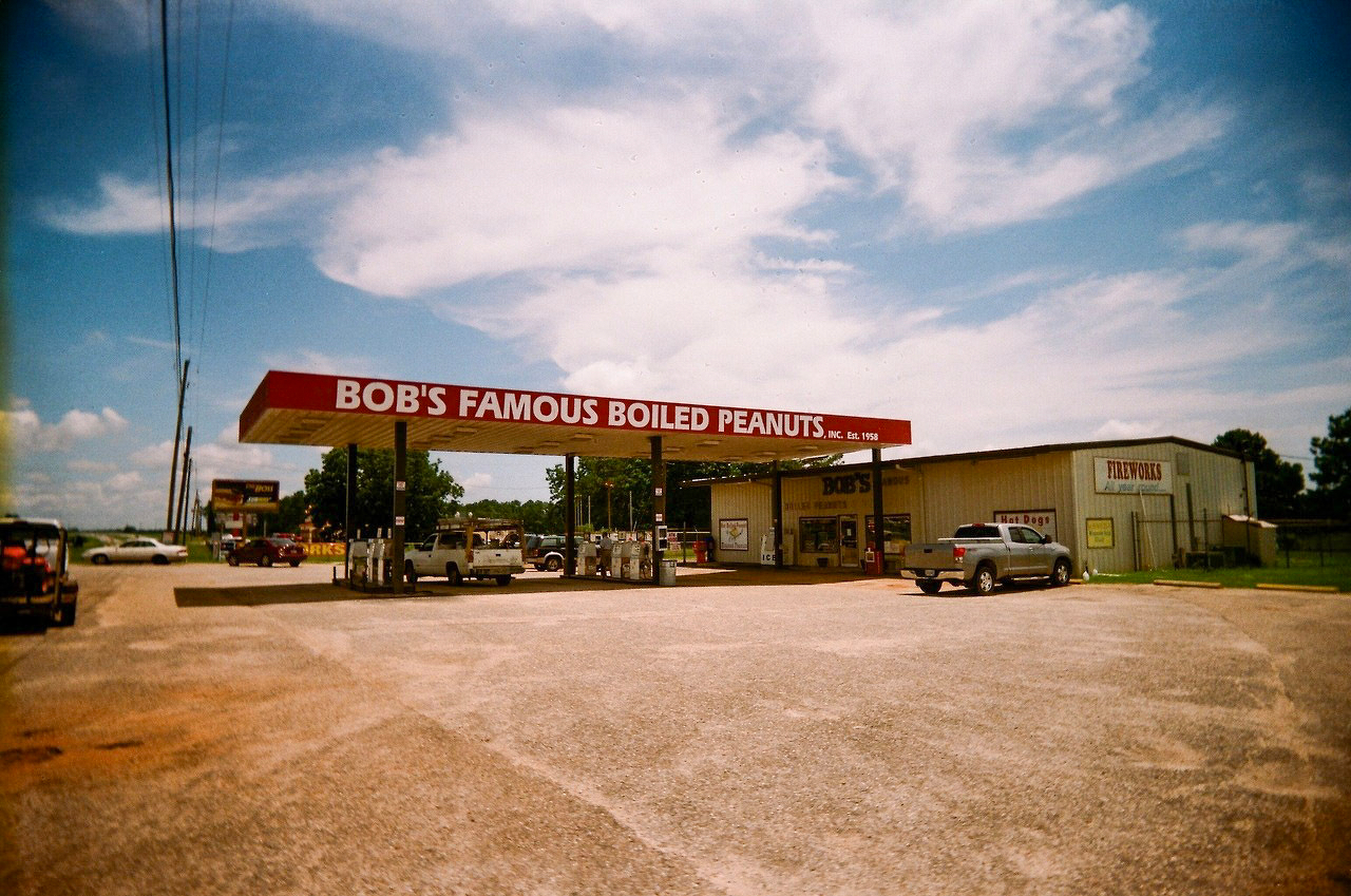 Auto-generated description: A roadside store with a sign for Bob&rsquo;s Famous Boiled Peanuts features a gas station and a parking lot with parked vehicles under a partly cloudy sky.