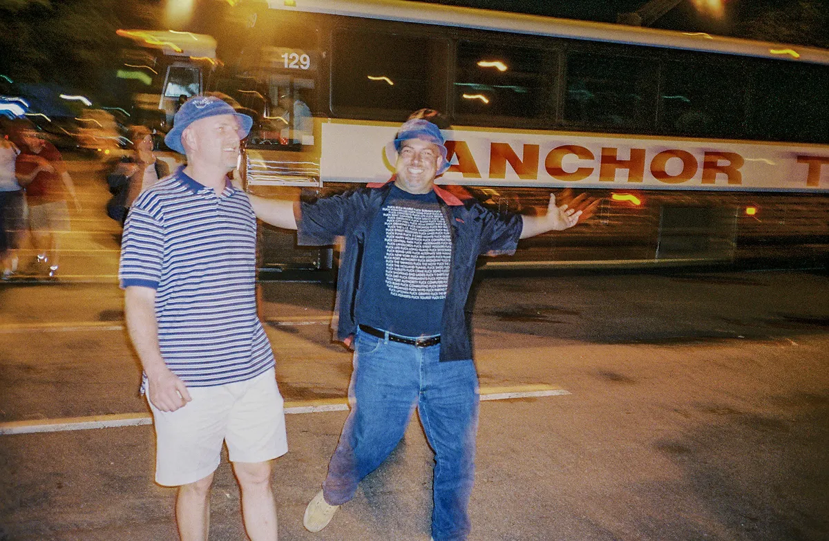 Auto-generated description: Two people wearing casual clothes and blue caps are posing happily on a street at night in front of a bus.
