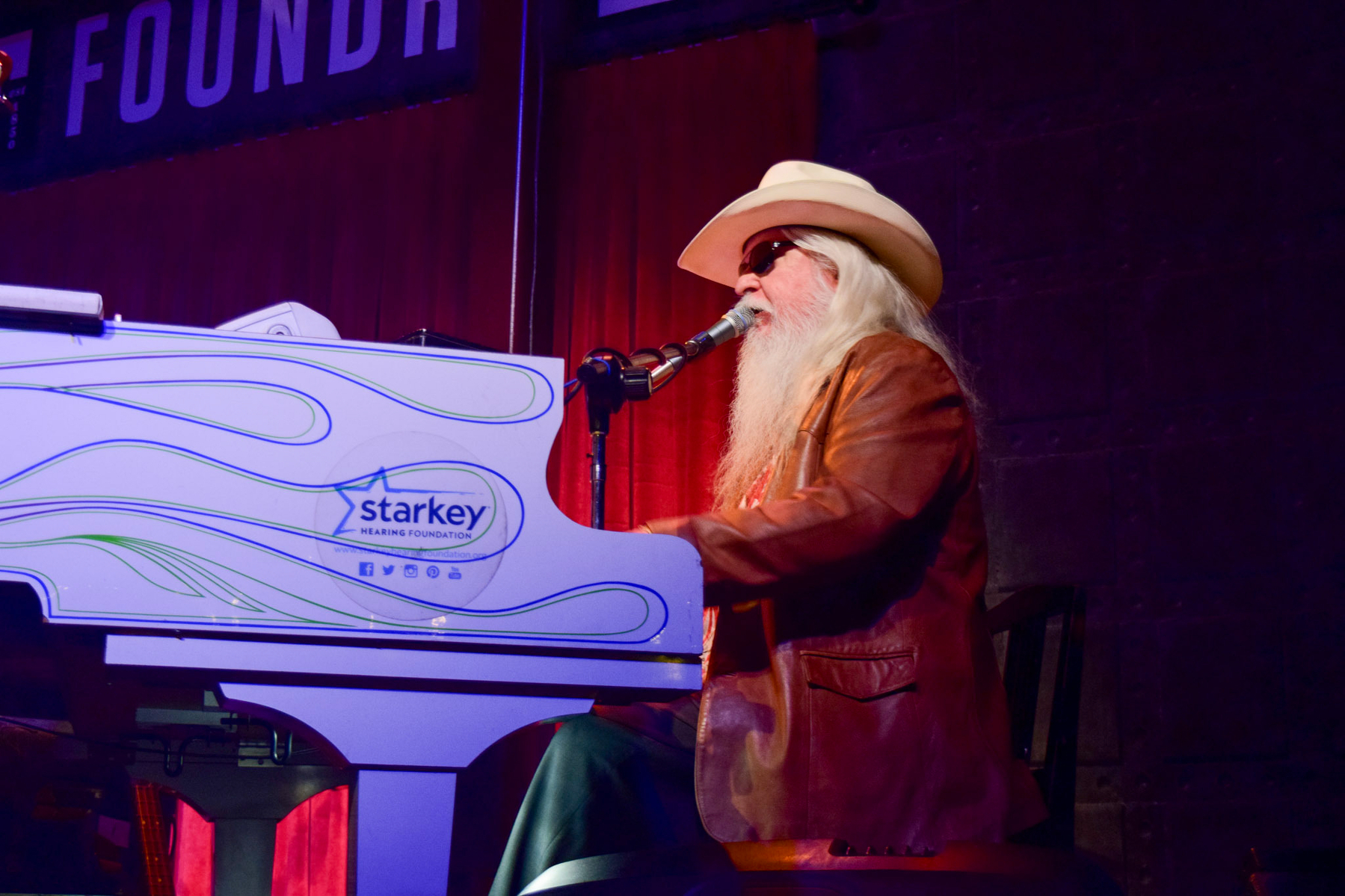 Auto-generated description: A musician wearing a cowboy hat and sunglasses plays the piano on stage under colorful lighting.