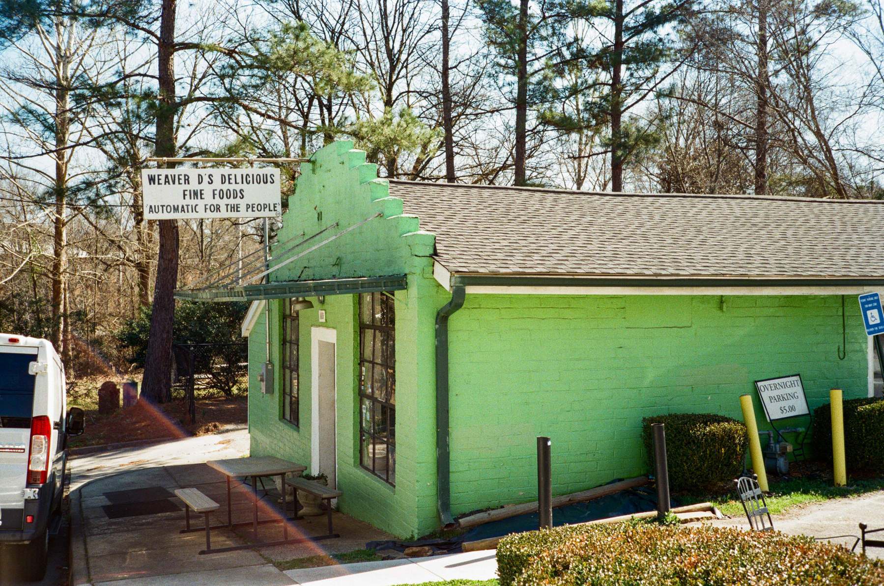 Auto-generated description: A green building with a sign reading Weaver D&rsquo;s Delicious Fine Foods is surrounded by trees and a parking area.