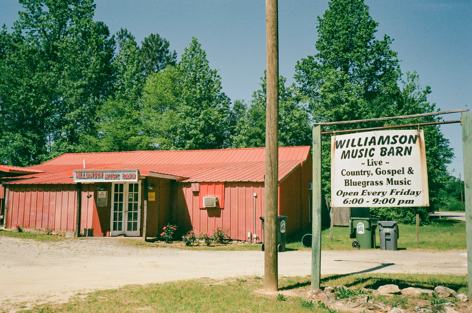 Auto-generated description: A rustic building with a red roof is situated next to a sign for Williamson Music Barn, advertising live country, gospel, and bluegrass music performances every Friday.
