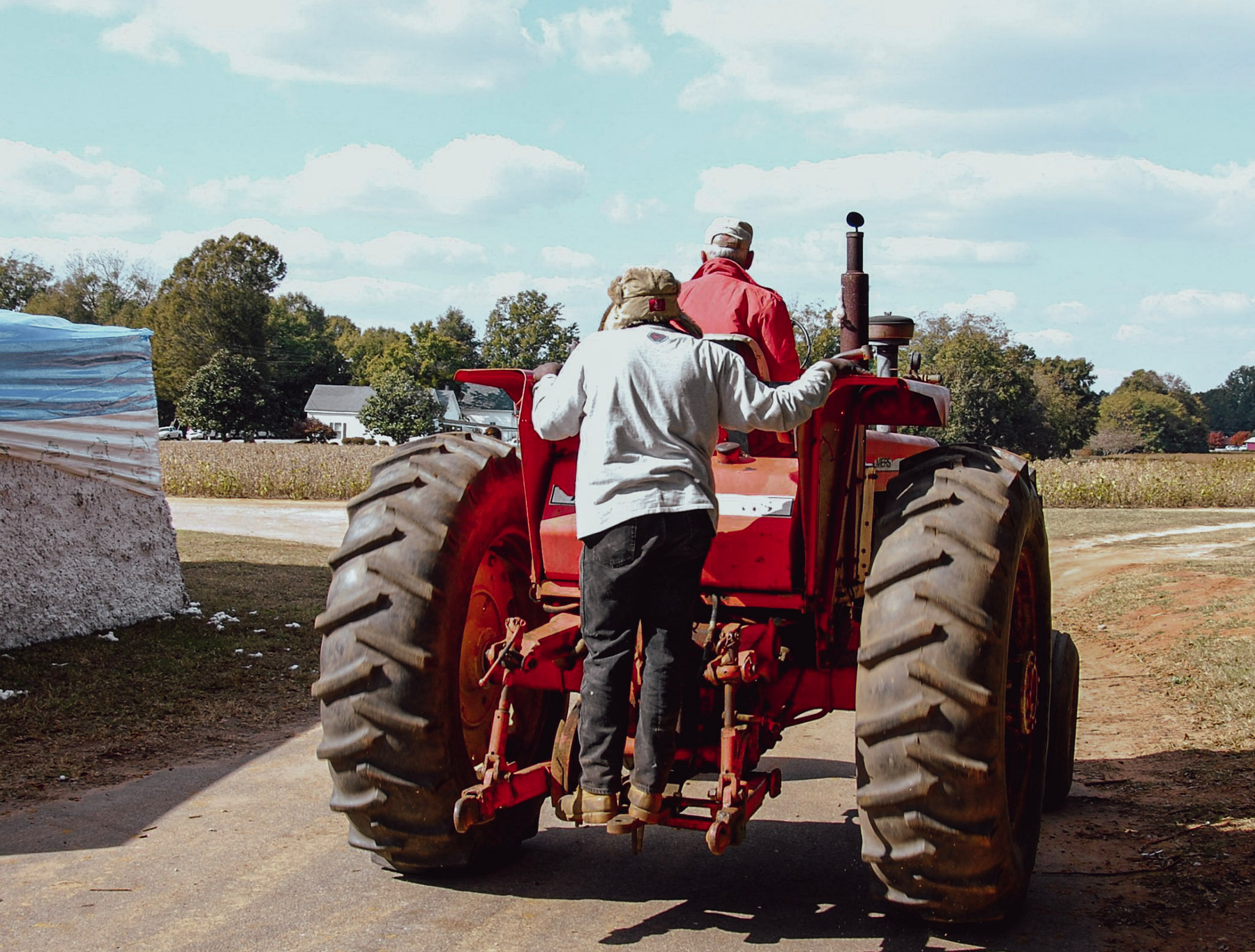 Auto-generated description: Two people, one standing on a red tractor and the other driving, are traveling on a rural road with farm fields in the background.