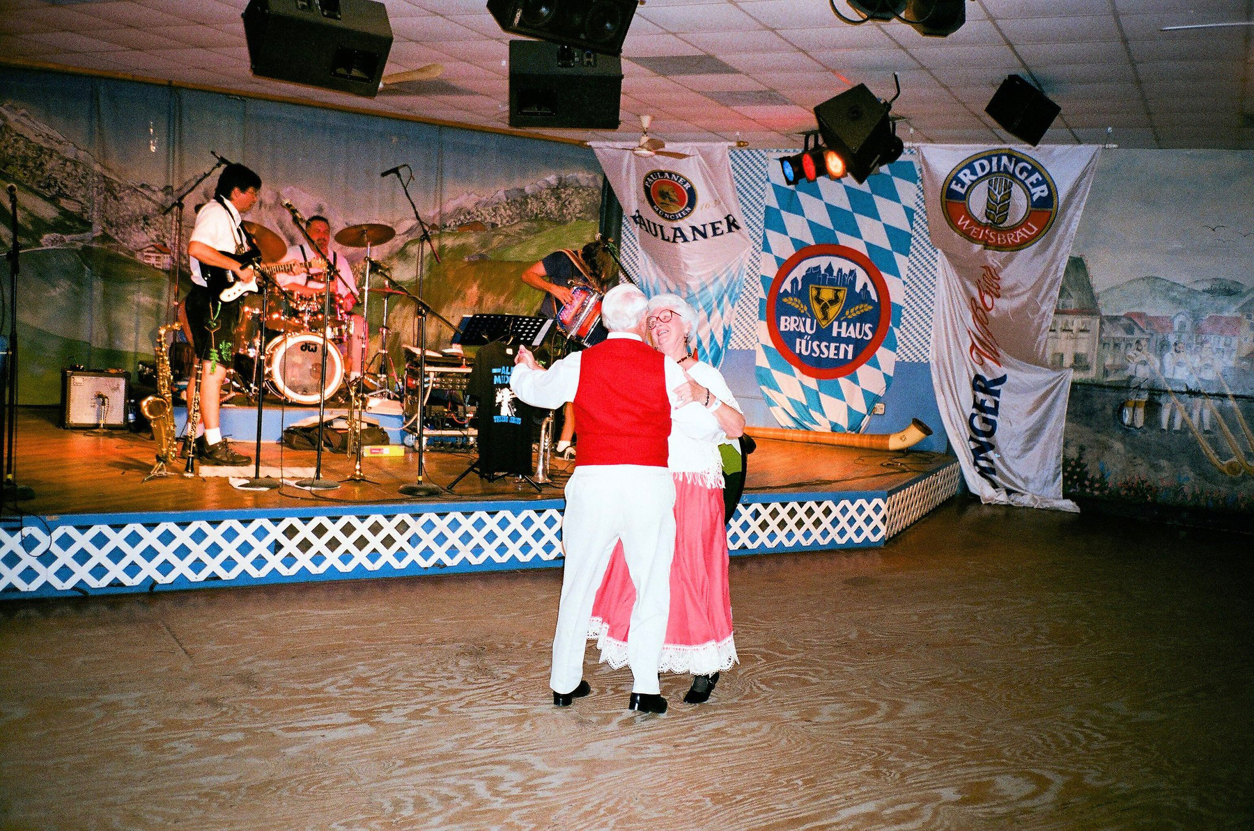 Auto-generated description: A couple in traditional Bavarian attire dances in front of a band performing on stage in a venue decorated with beer festival banners.