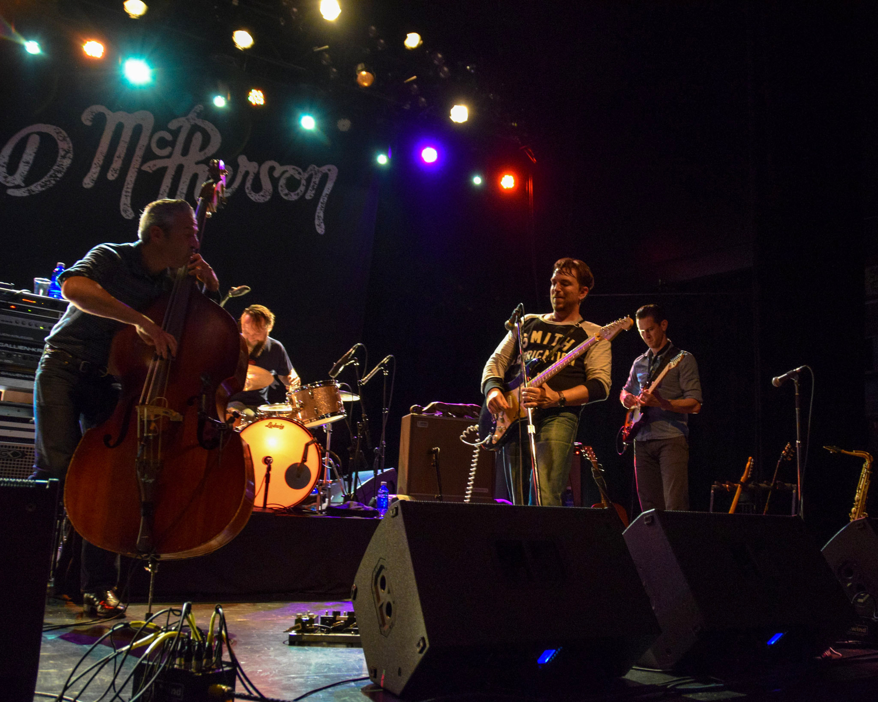 Auto-generated description: A band is performing on stage with a bassist, drummer, guitarist, and saxophonist under vibrant stage lights.