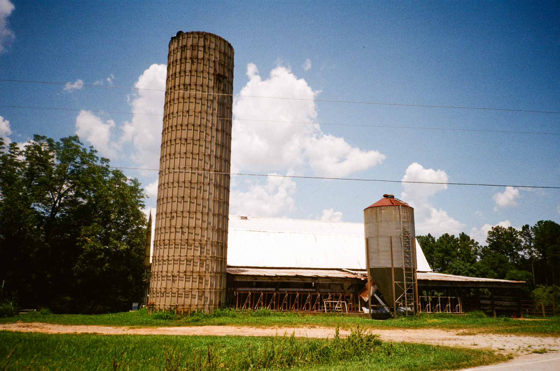 Auto-generated description: A rural scene with a tall silo, a barn with a white roof, and a smaller red and white silo, set against a backdrop of trees and a partly cloudy sky.