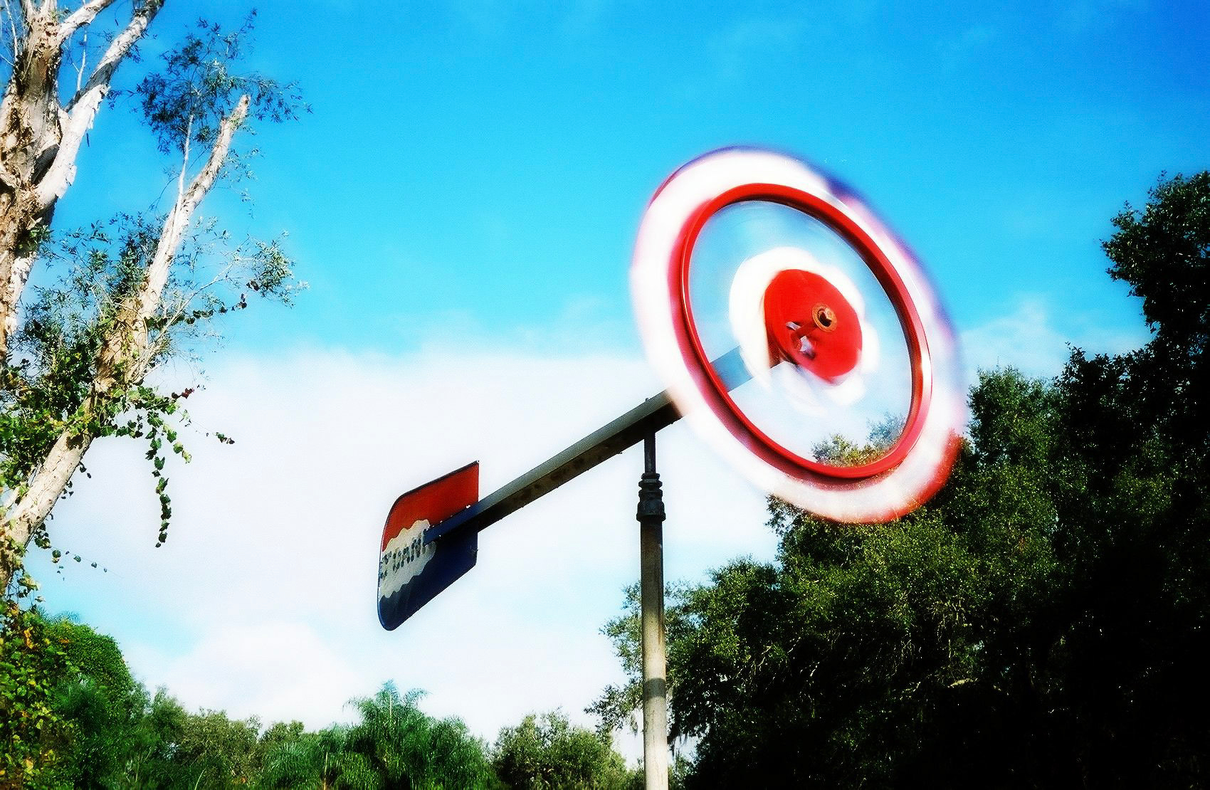Auto-generated description: A spinning circular sign on a pole with Pepsi branding is set against a backdrop of trees and a blue sky.