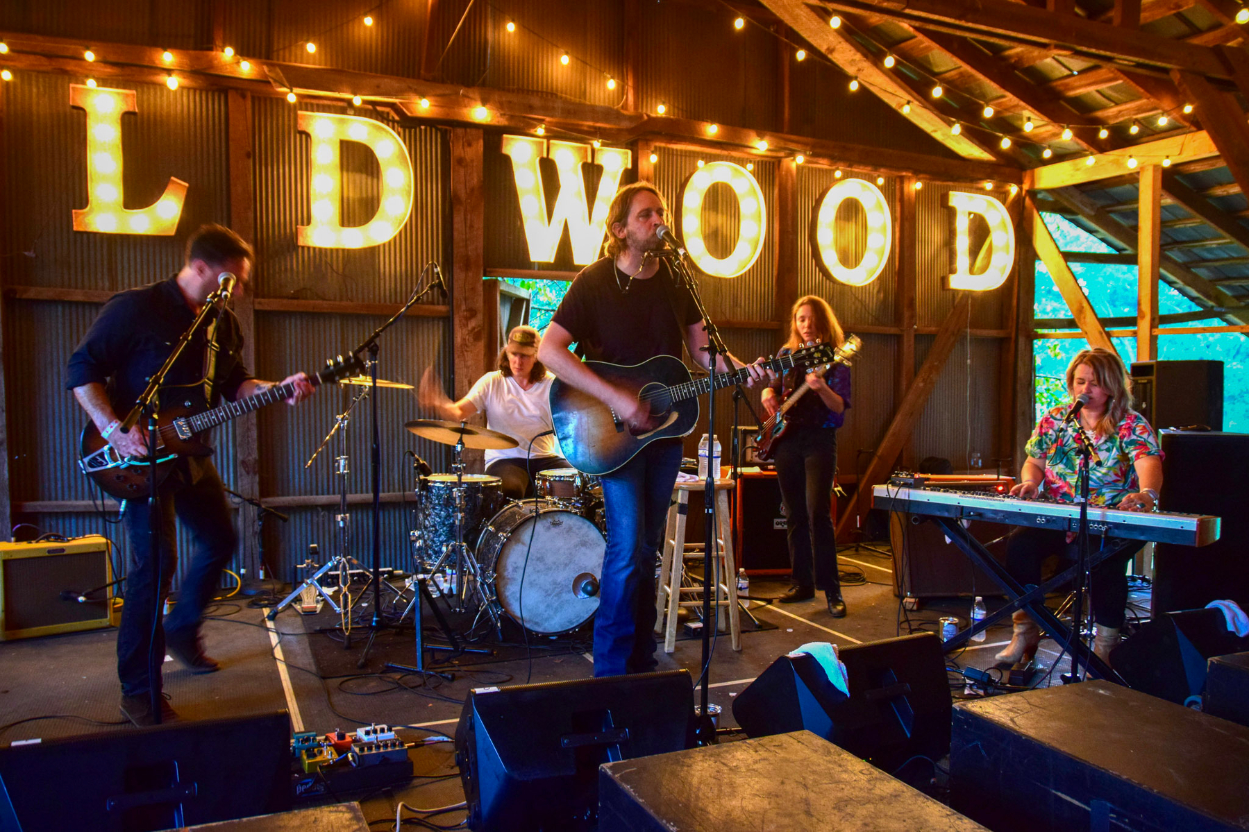Auto-generated description: A band is performing on stage with a lit WILDWOOD sign in the background and string lights overhead.