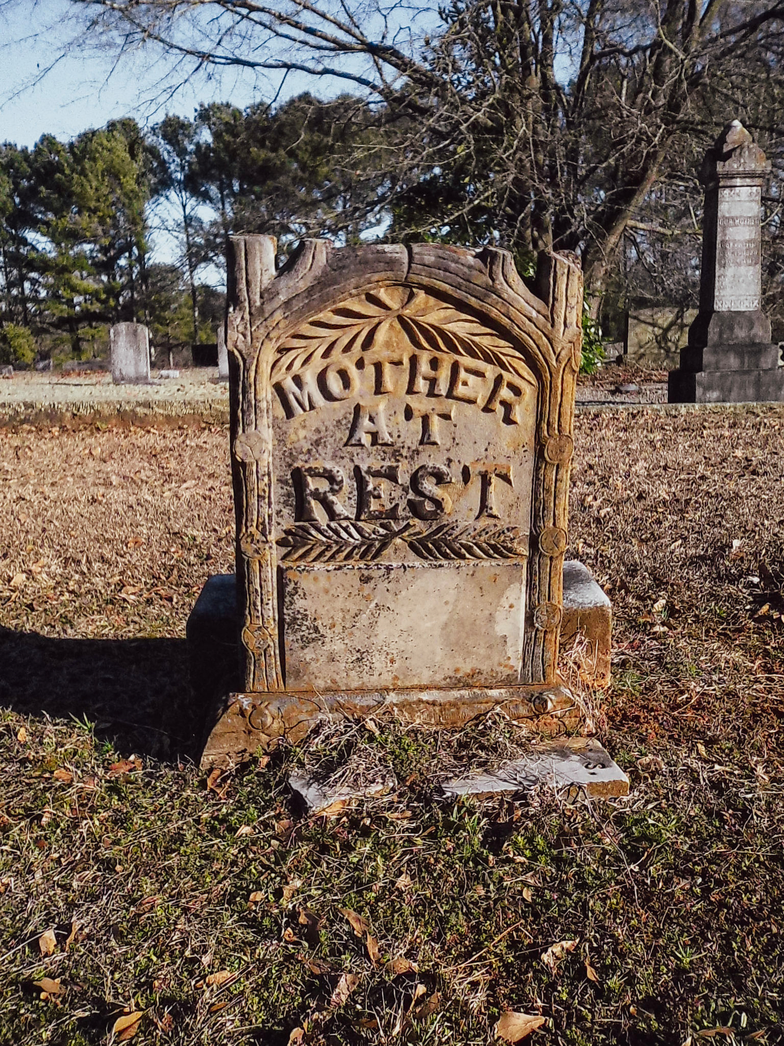 Auto-generated description: A weathered gravestone with intricate carvings reads MOTHER AT REST amidst a peaceful cemetery backdrop.