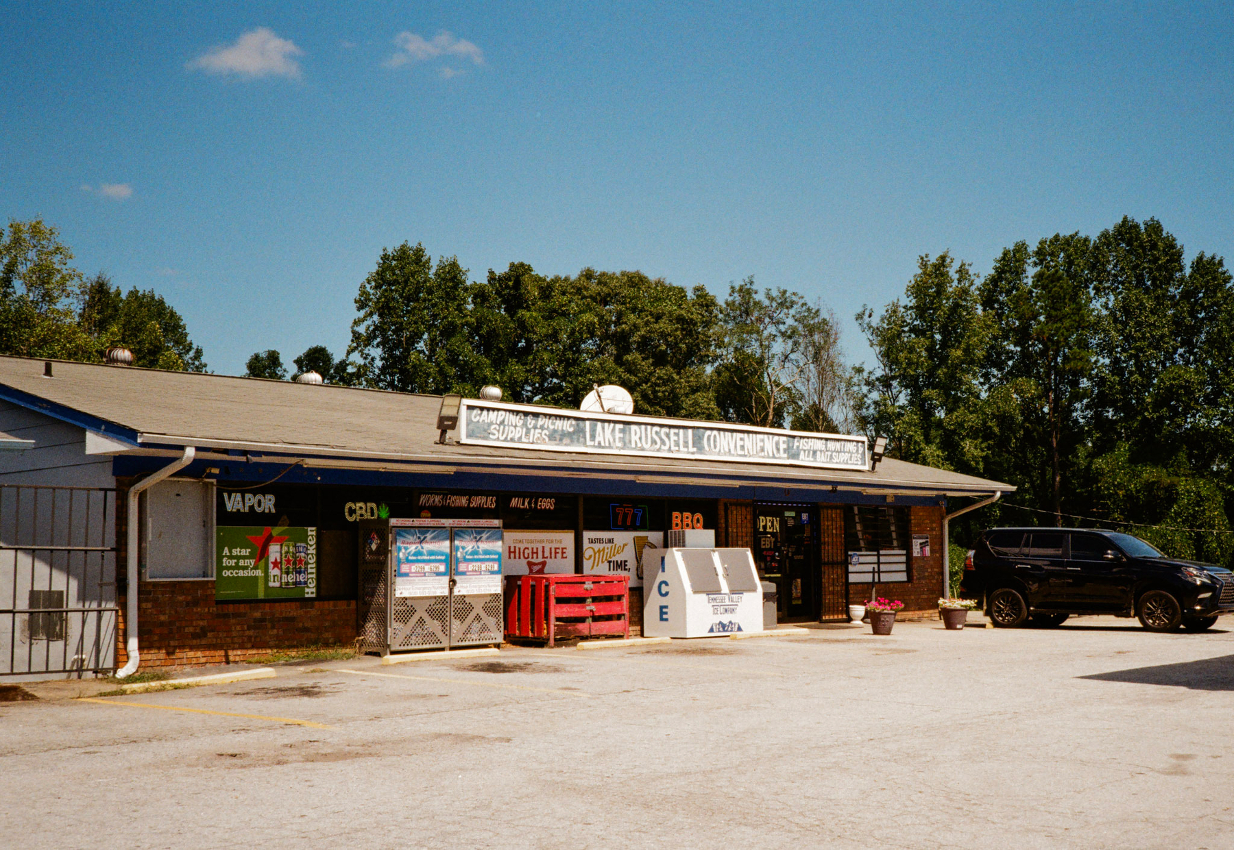 Auto-generated description: A small convenience store with a sign labeled Lake Russell Convenience features advertisements for various products, including ice and BBQ, with a car parked in front.