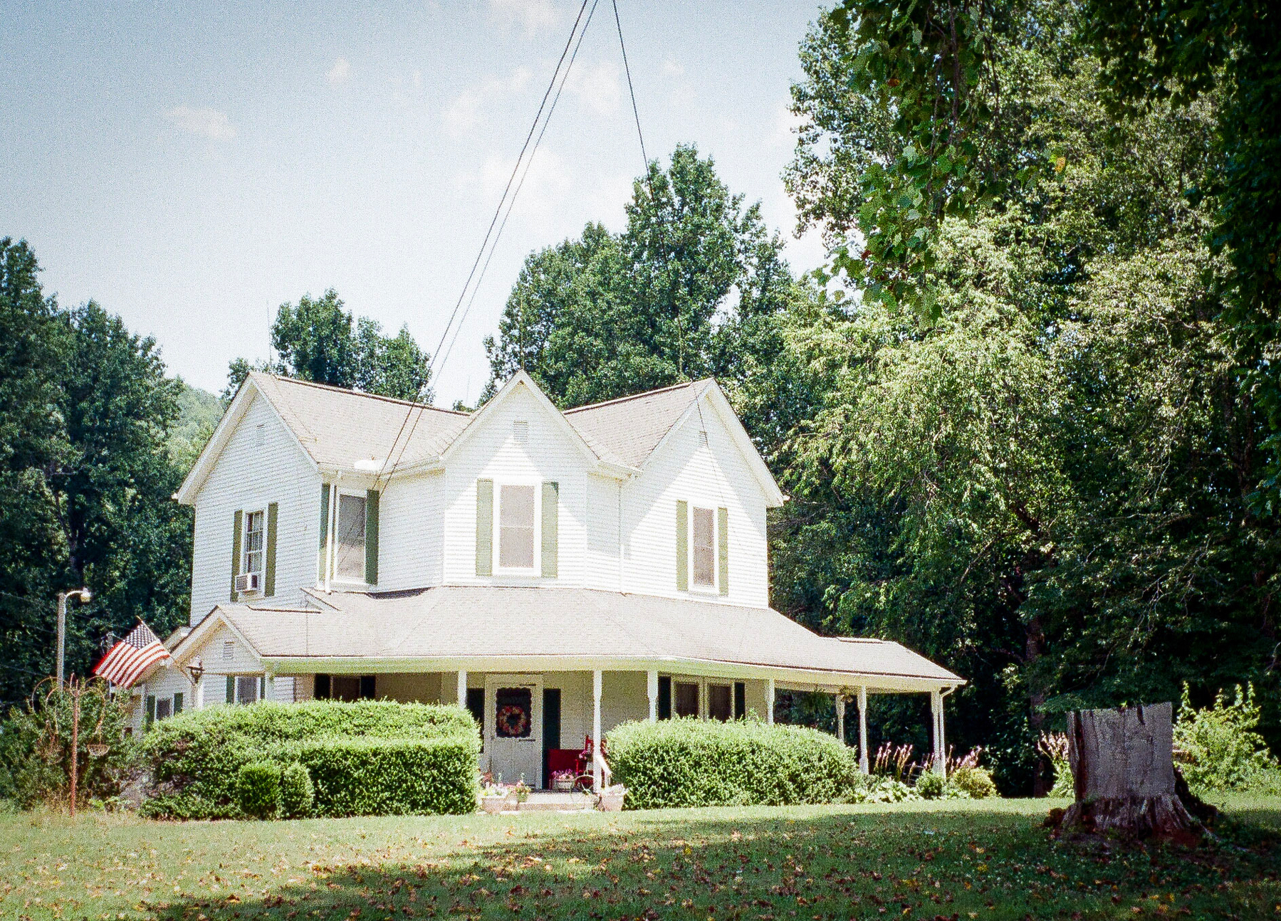 Auto-generated description: A white two-story house with a porch and surrounded by greenery sits in a scenic yard with a flagpole and a tree stump in the foreground.