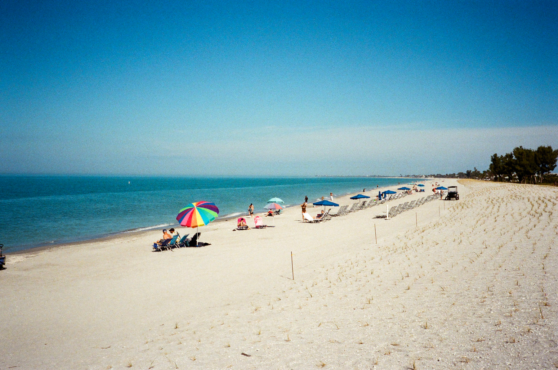 Auto-generated description: A sandy beach features several people relaxing under colorful umbrellas near the ocean on a clear, sunny day.