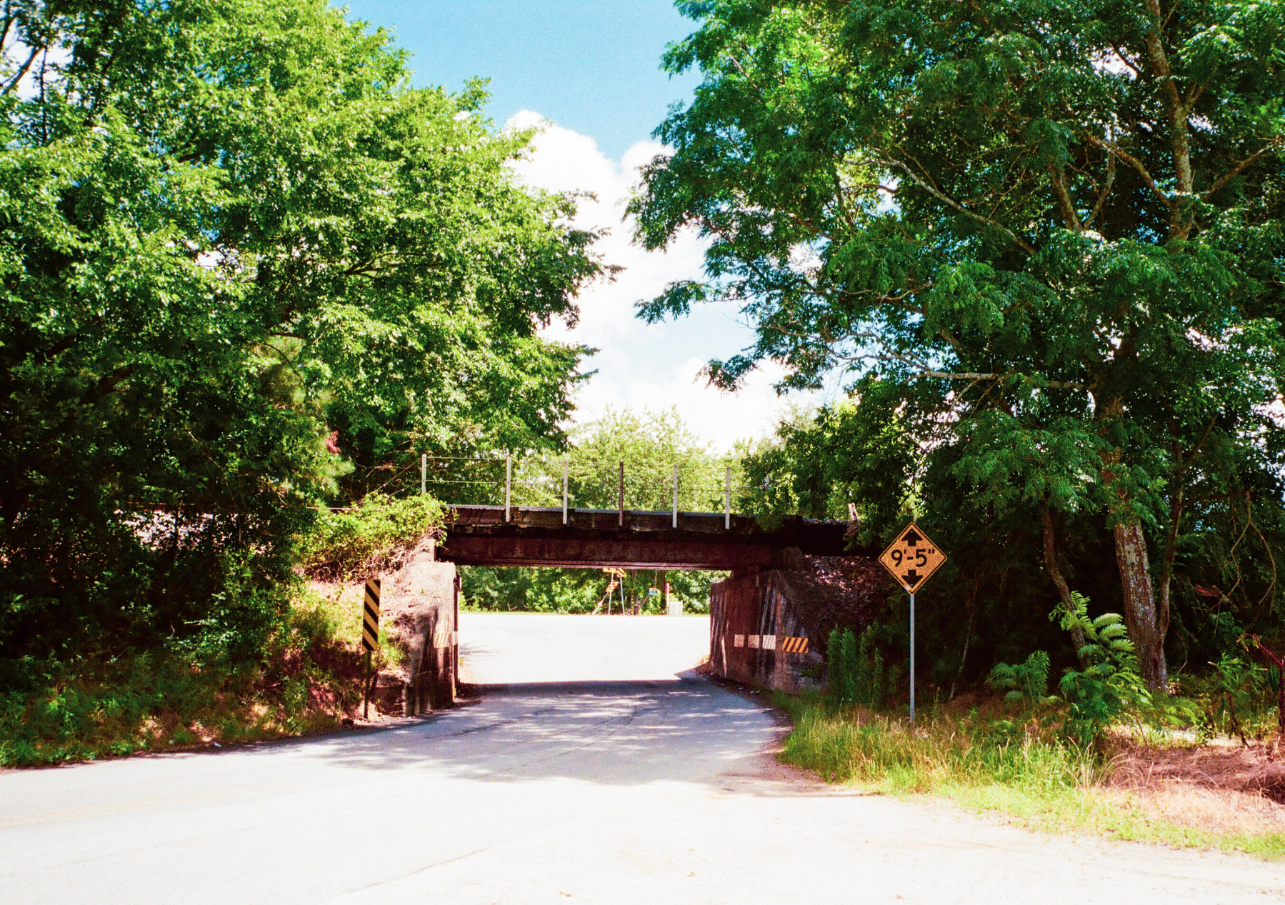 Auto-generated description: A road passes under a small bridge surrounded by lush green trees, with a height restriction sign visible on the right.