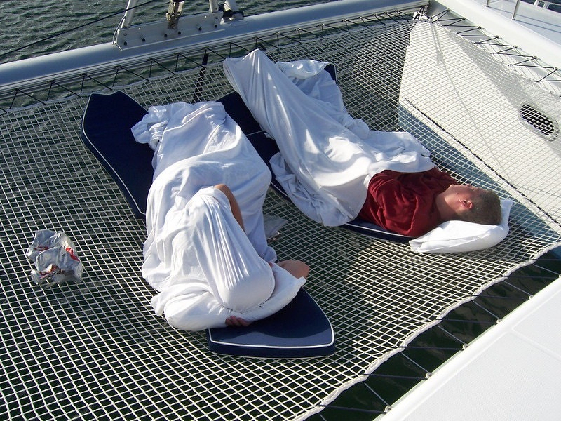 Auto-generated description: Two people are sleeping on sun loungers covered with blankets on a netting area of a boat.