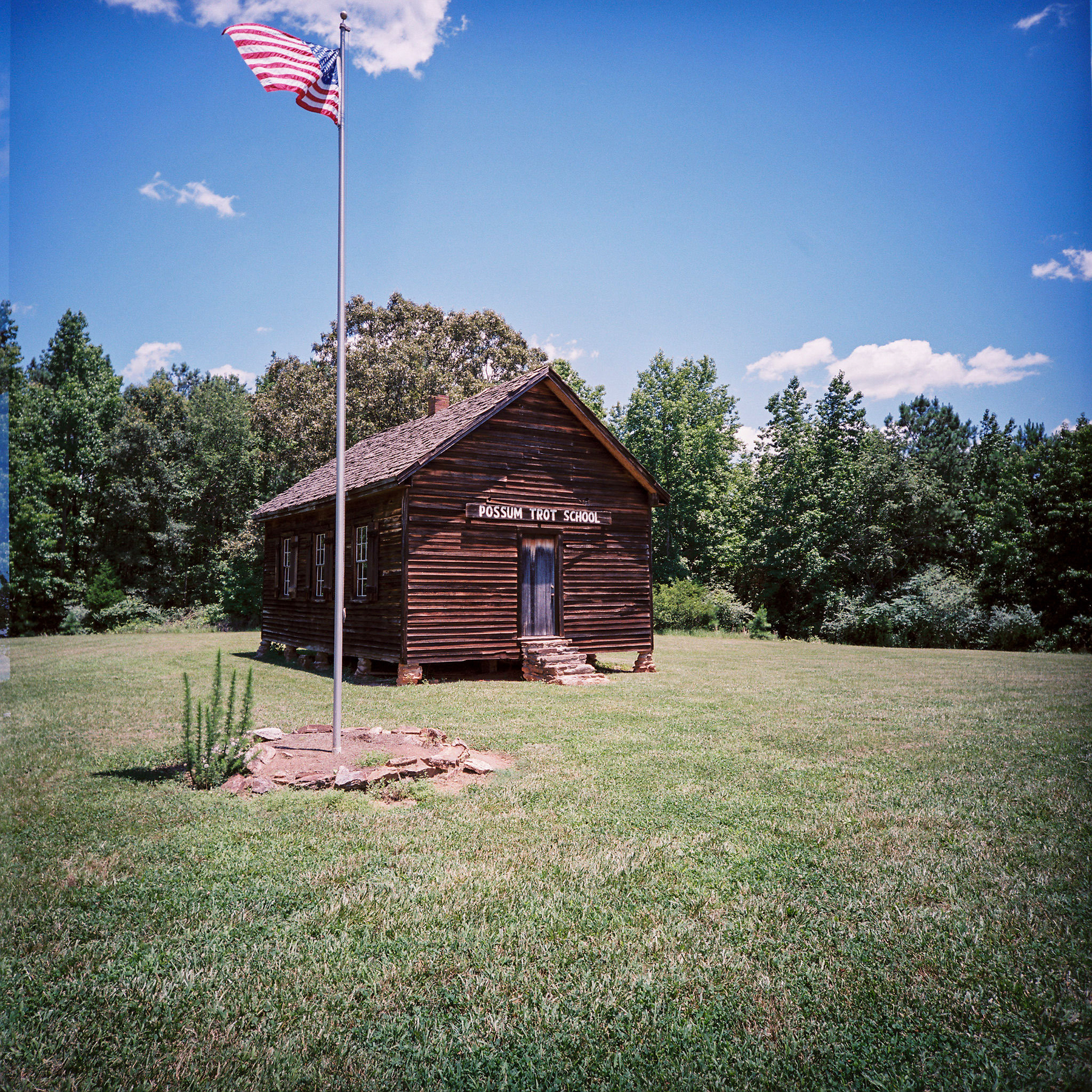 Auto-generated description: A rustic wooden building labeled Possum Trot School is set in a grassy area with an American flag flying in front.