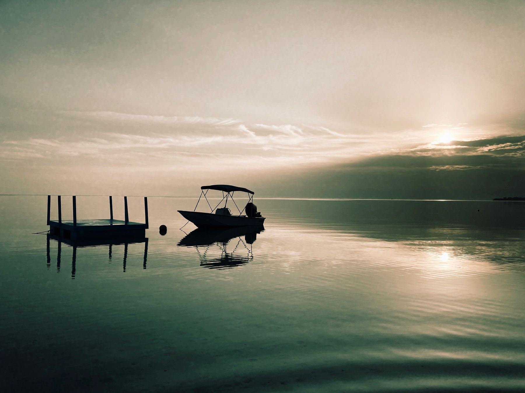 Auto-generated description: A tranquil scene features a small boat on calm waters with a floating platform, under a sky with the sun partially obscured by clouds.