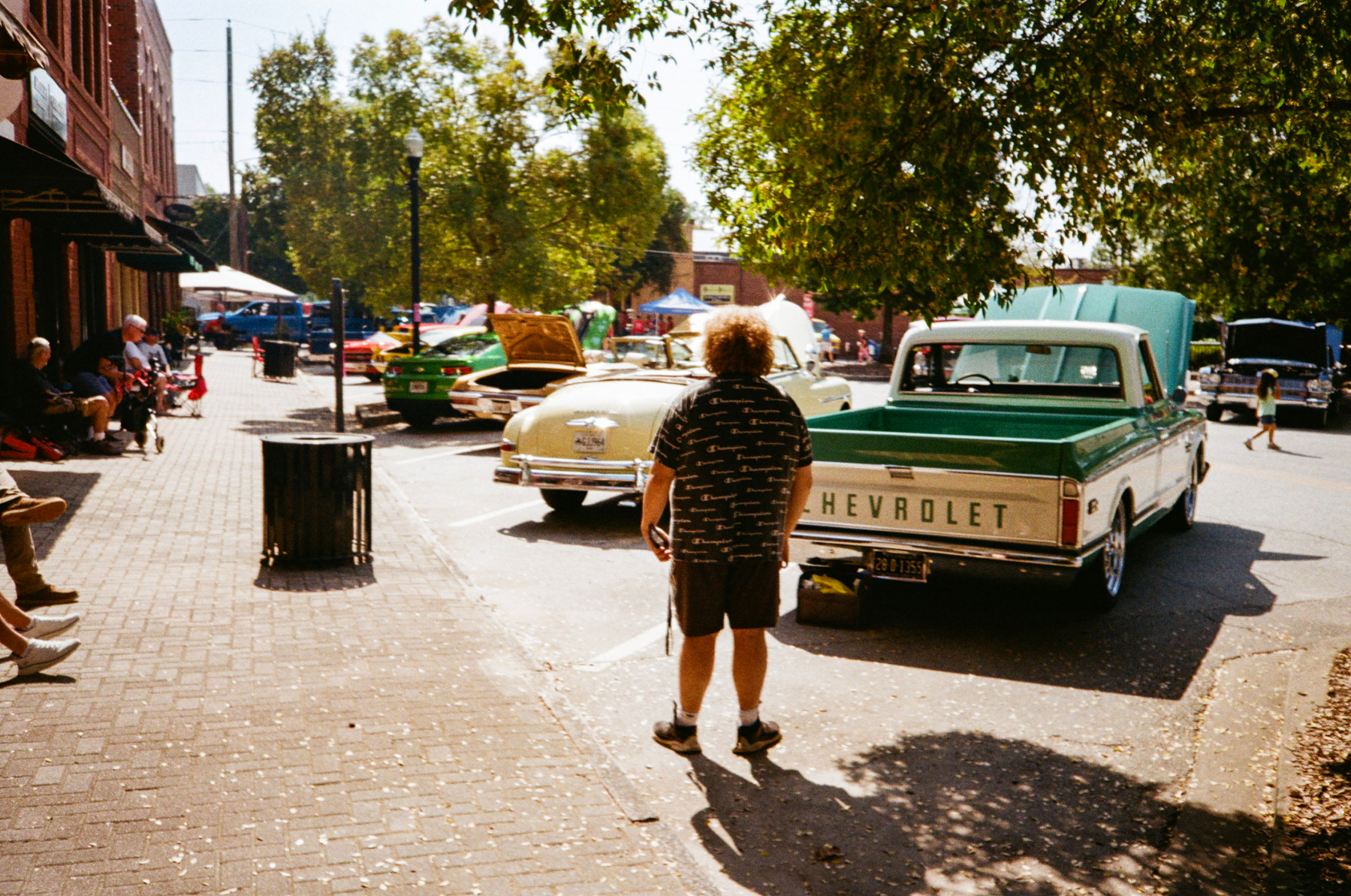 Auto-generated description: A person stands on a brick-paved street near classic cars, including a Chevrolet, under the shade of trees.