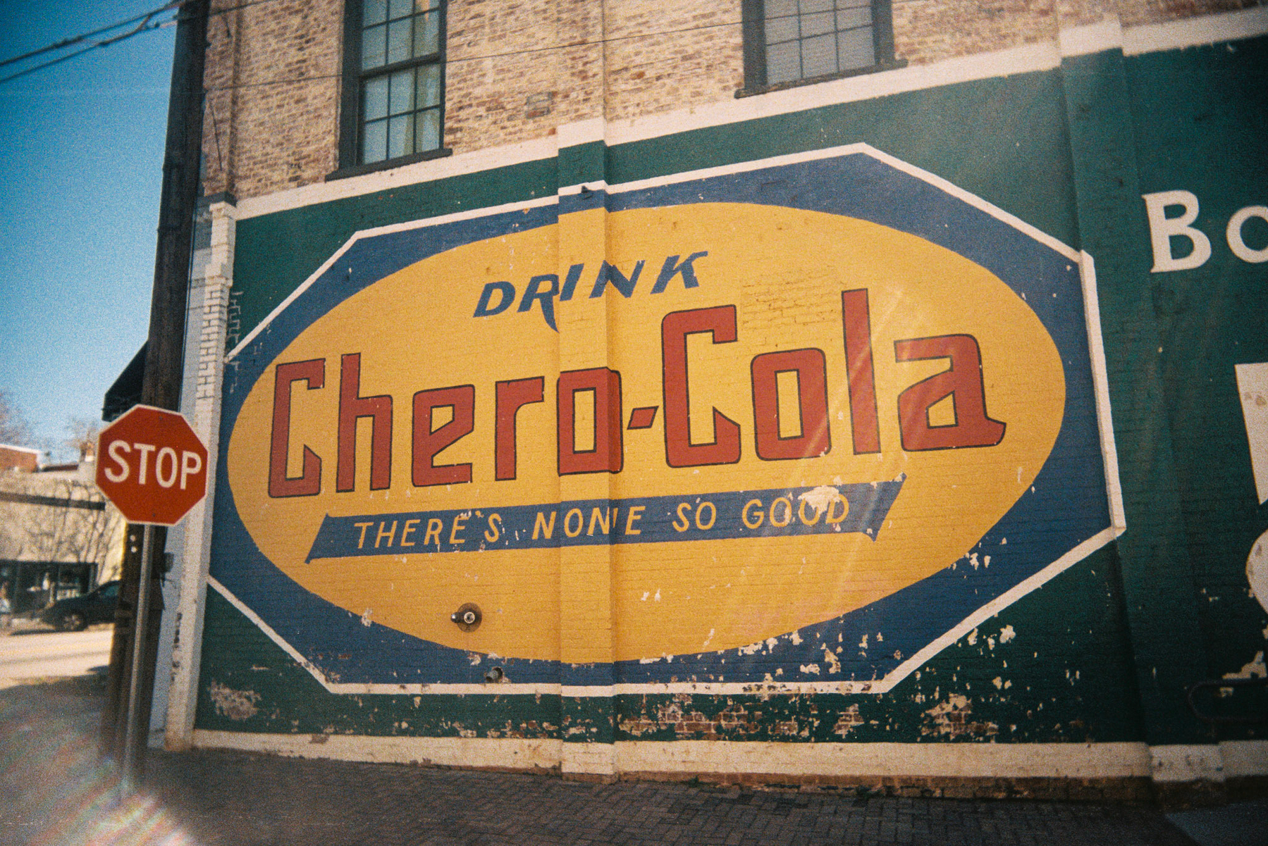 Auto-generated description: A vintage mural advertises Drink Chero-Cola with the slogan There&rsquo;s None So Good painted on a brick wall next to a stop sign.