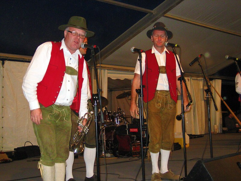 Auto-generated description: Two men wearing traditional Bavarian outfits stand on a stage with musical instruments and microphones.