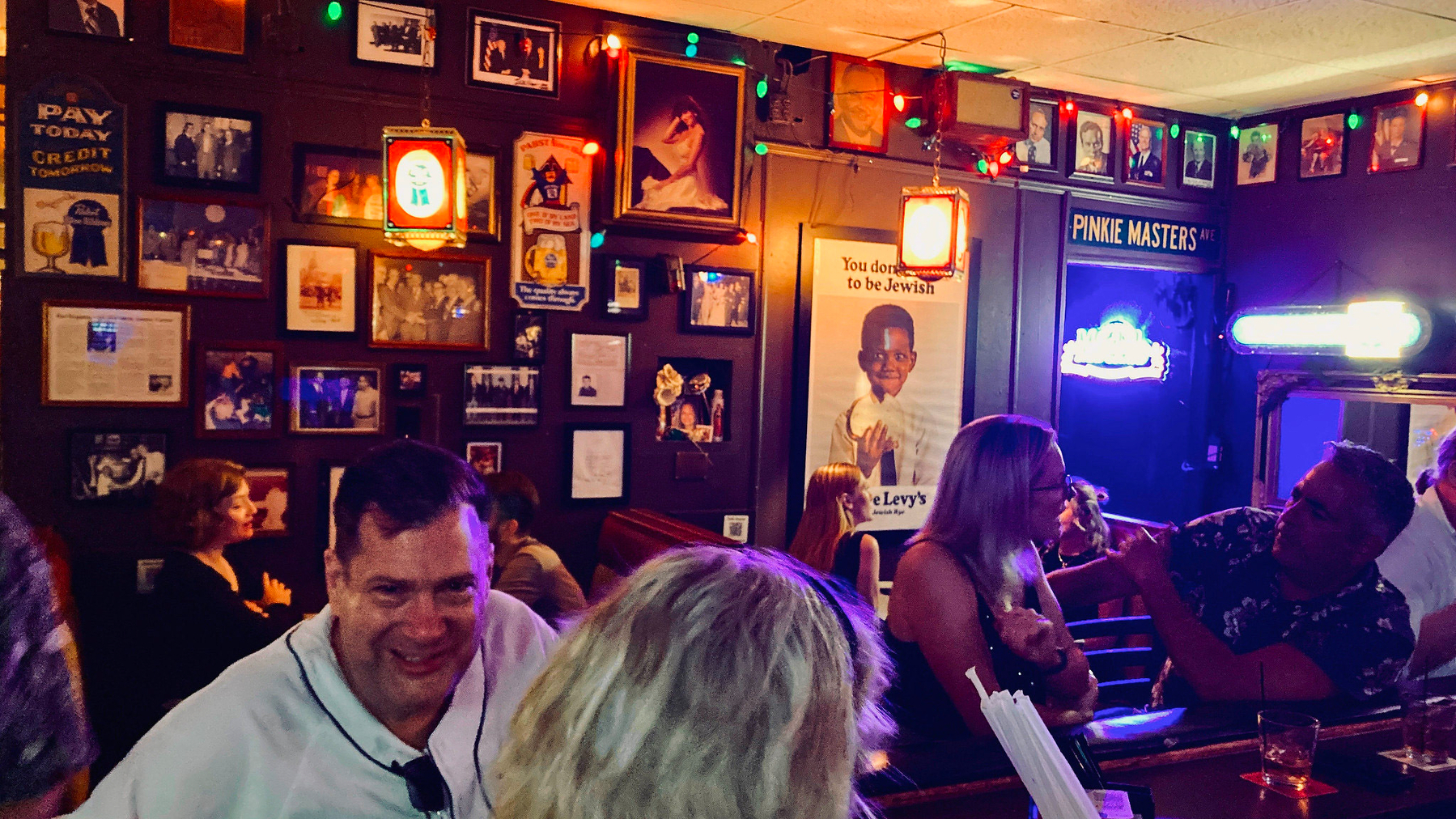 Auto-generated description: A cozy bar is filled with people enjoying themselves, surrounded by colorful lights and various framed pictures on the walls.