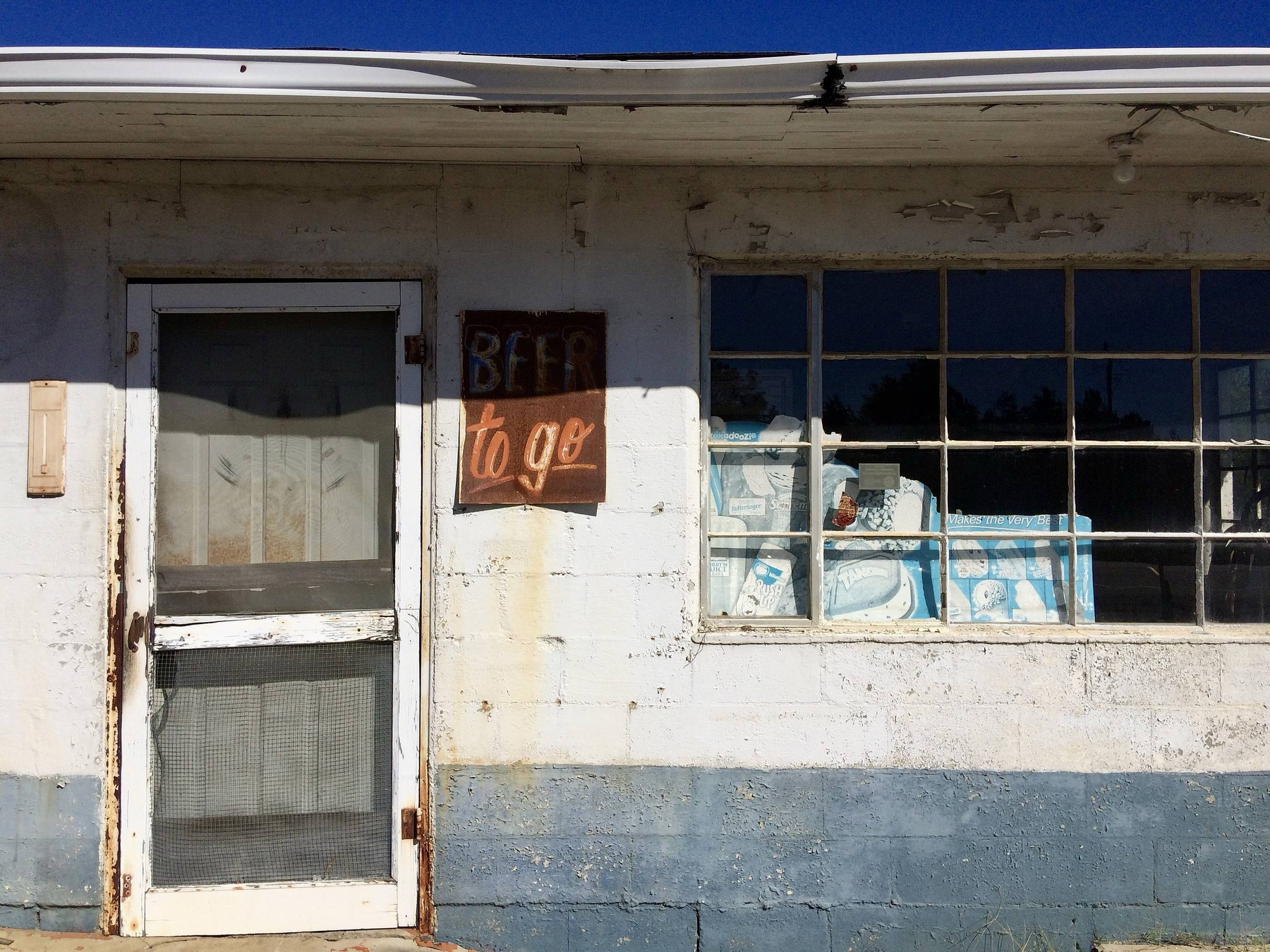 Auto-generated description: A weathered storefront displays a Beer to go sign next to a window filled with cases of drinks.